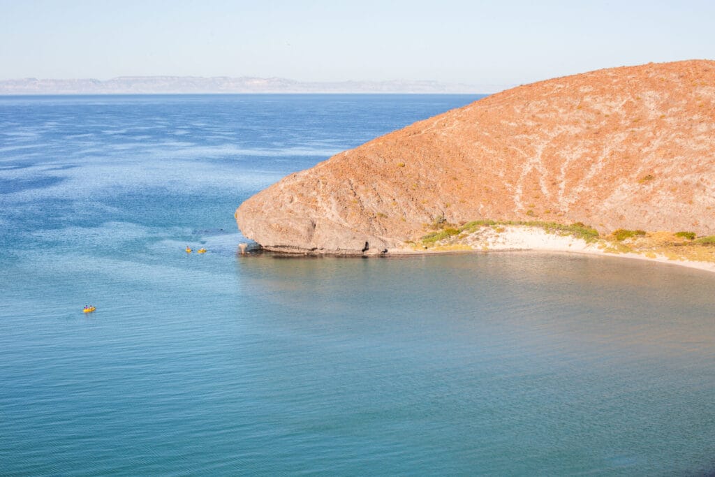 Calm blue sea next to a rocky hill, with kayakers exploring the coastline. Clear sky above adds to the serene landscape.