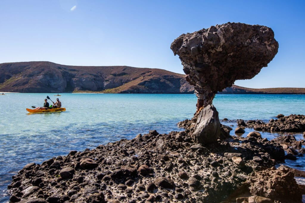 Two people kayak near a Mushroom Rock formation in clear, turquoise water. The background features rugged, brown hills under a clear blue sky.