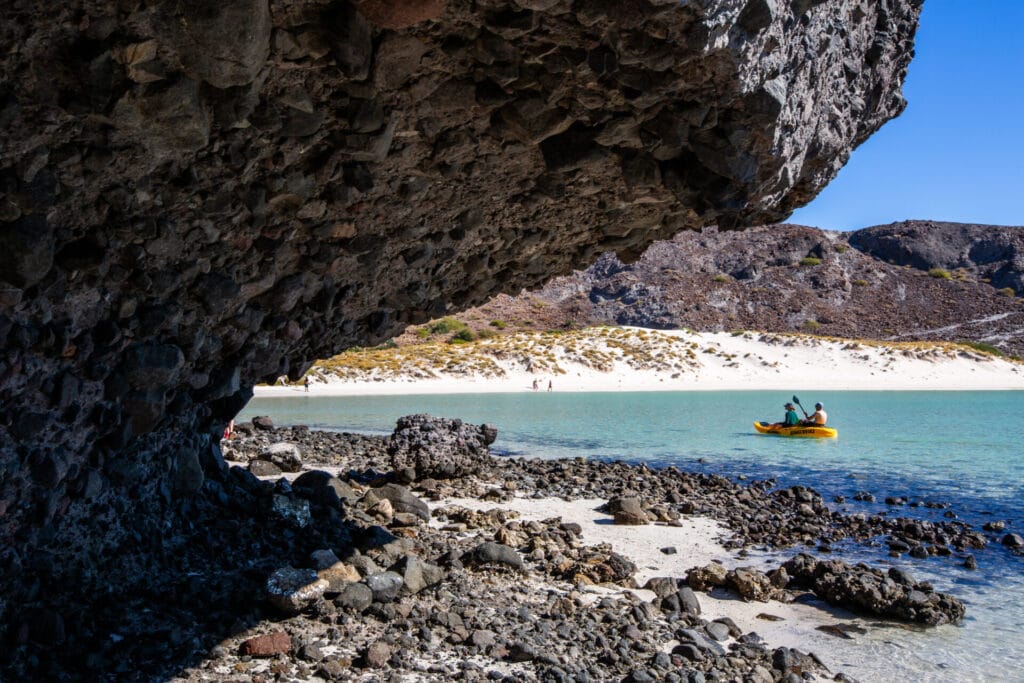A rocky coastal scene features a person kayaking on turquoise waters, sandy beach, and overhanging rock formation under a clear blue sky.