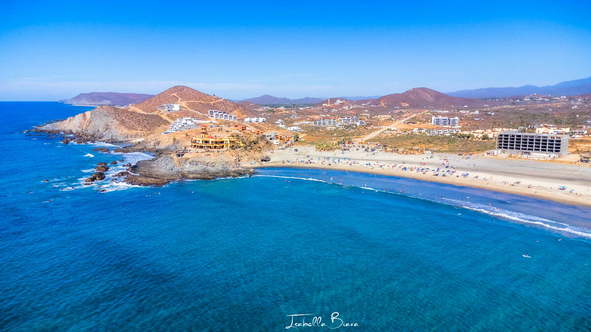 Coastal landscape with a sandy beach, clear blue water, rocky hills, scattered buildings, and people enjoying the shore. Clear skies enhance the scenic view.