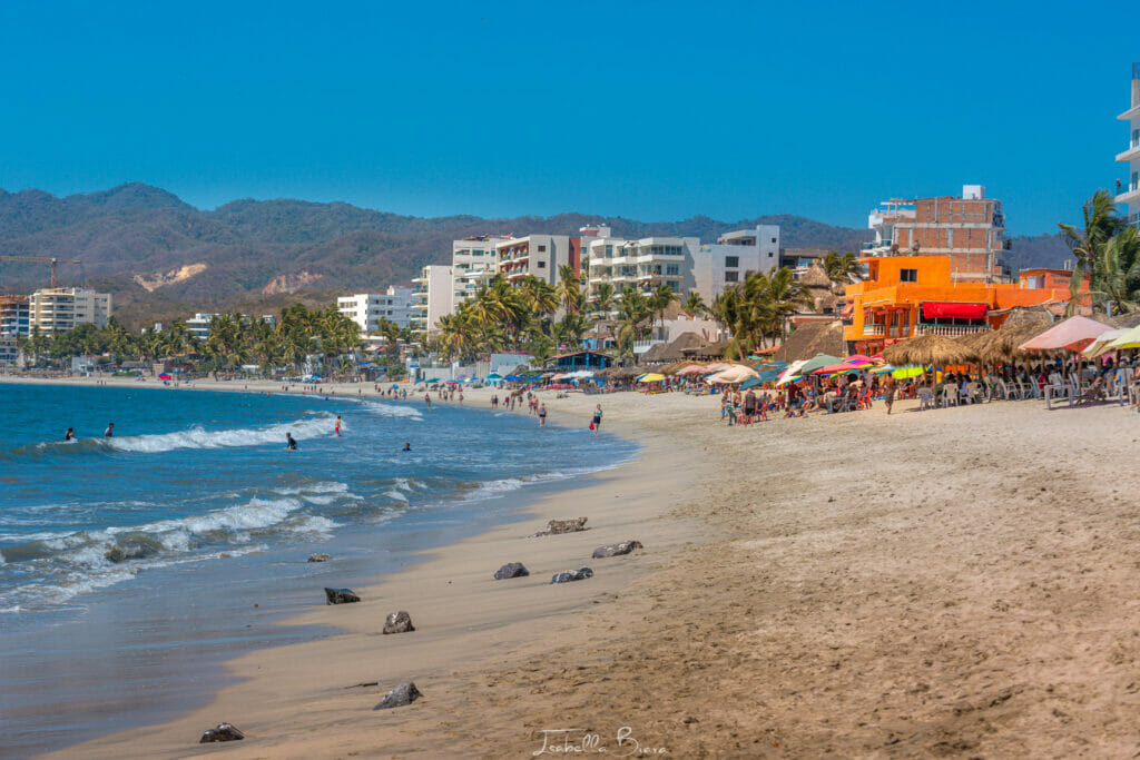 Wide sandy beach in Bucerías filled with colorful umbrellas, sunbathers, and gentle surf, with modern beachfront buildings and mountains in the distance.