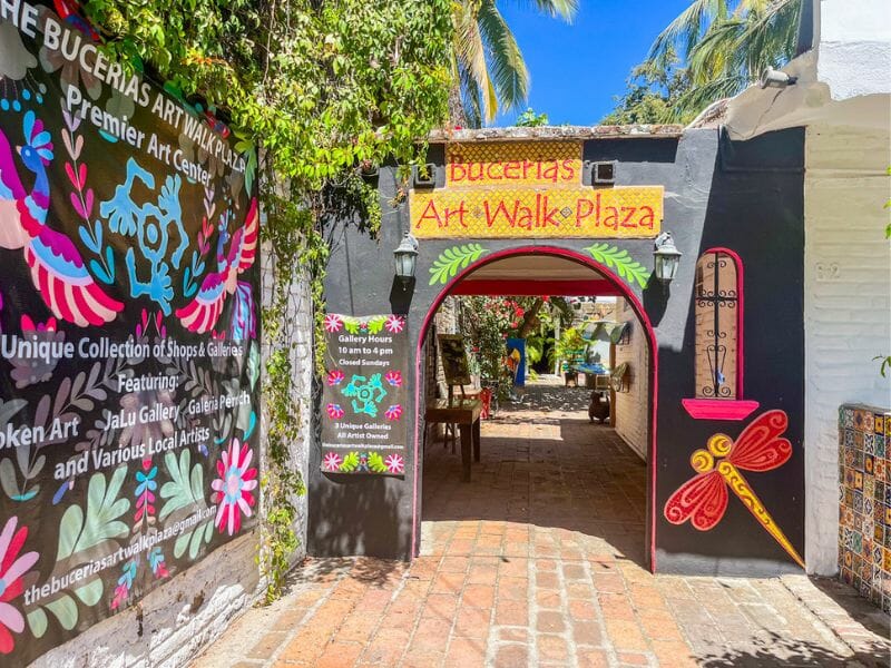 Bright entrance to Bucerías Art Walk Plaza with hand-painted signs and murals featuring flowers, dragonflies, and gallery info, leading into a charming brick path courtyard.
