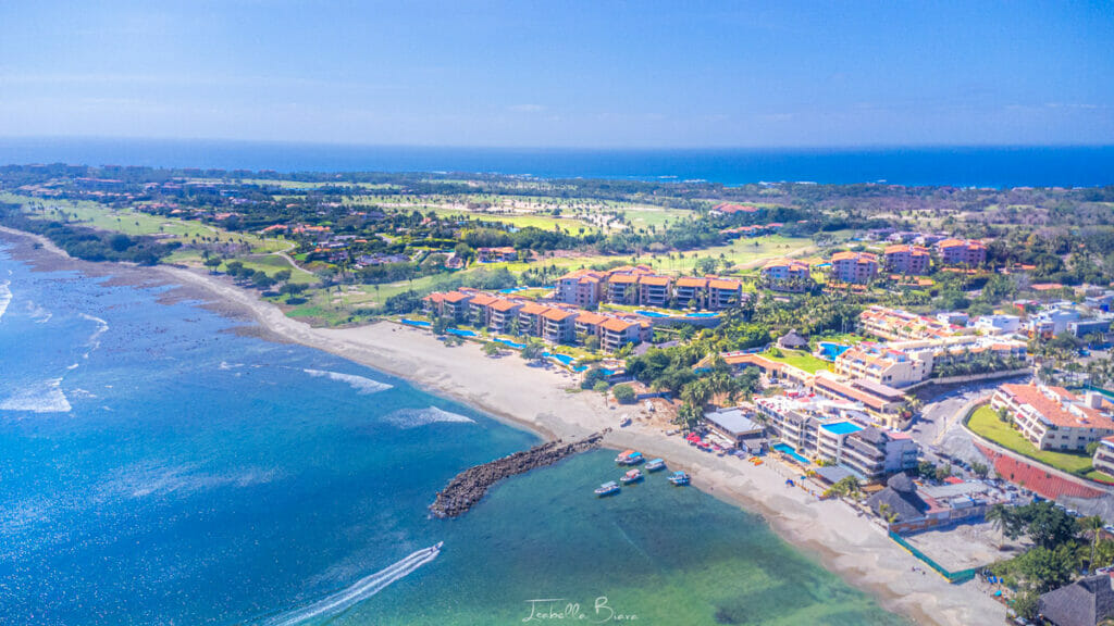 Drone view of Punta de Mita’s coastline, with luxury resorts, golf courses, and boats docked along a calm bay under a clear blue sky.