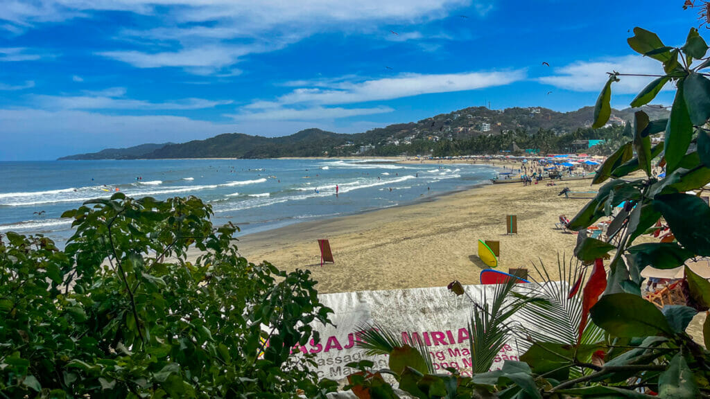 View of Sayulita beach from behind tropical greenery, showing a busy shoreline with beachgoers, umbrellas, and small waves rolling in under a bright sky
