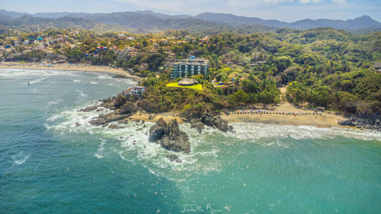 Drone shot showing Sayulita’s curving coastline, rocky point, and the popular Playa de los Muertos lined with umbrellas, surrounded by dense green hills.