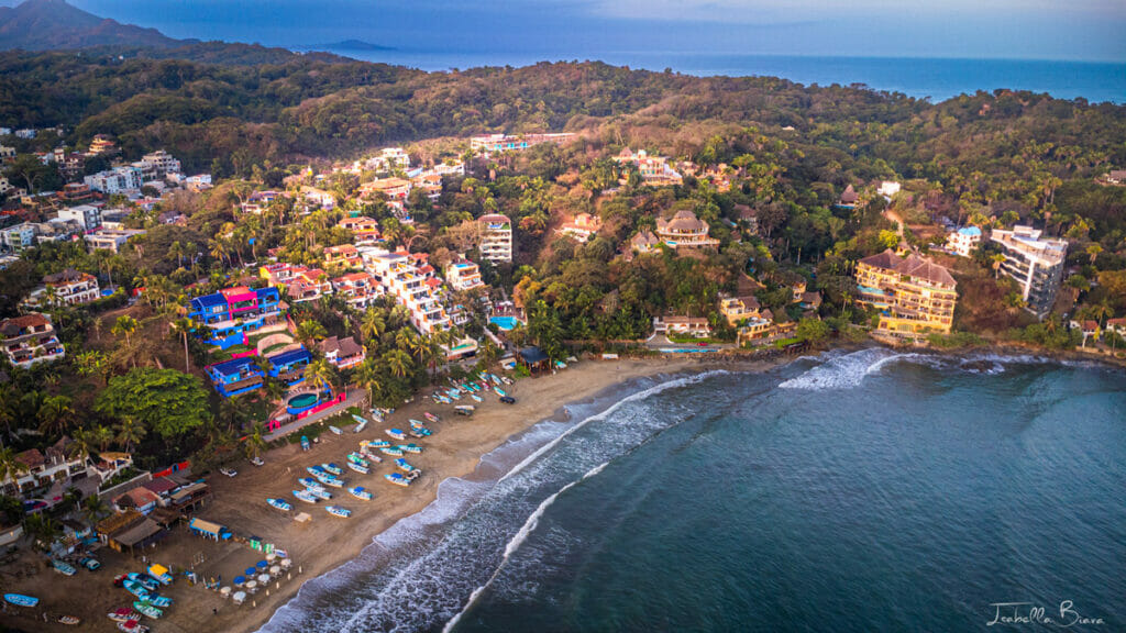 Aerial view of Sayulita's main beach with surfers in the waves, beach umbrellas lining the sand, and colorful hillside homes backing the bay.