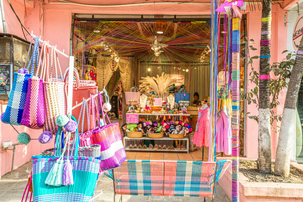 Vibrant storefront bursting with color, with rainbow woven bags, handmade dolls, and textiles hanging in and around the entrance of a small pink boutique.