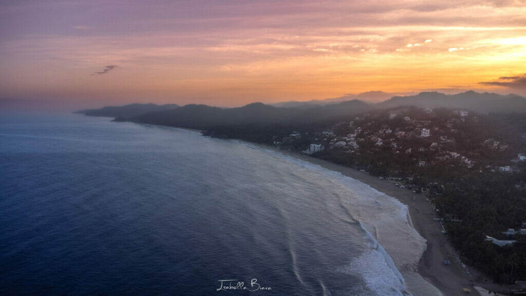 Aerial view of Sayulita's coastline at dawn, with the soft glow of a golden-orange sunrise washing over the ocean and silhouetted hills in the distance.
