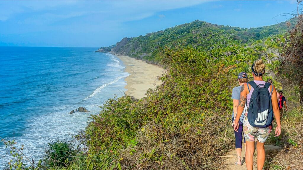 Scenic coastal hiking trail with people walking toward a secluded sandy beach below, surrounded by lush greenery and overlooking the blue Pacific Ocean.