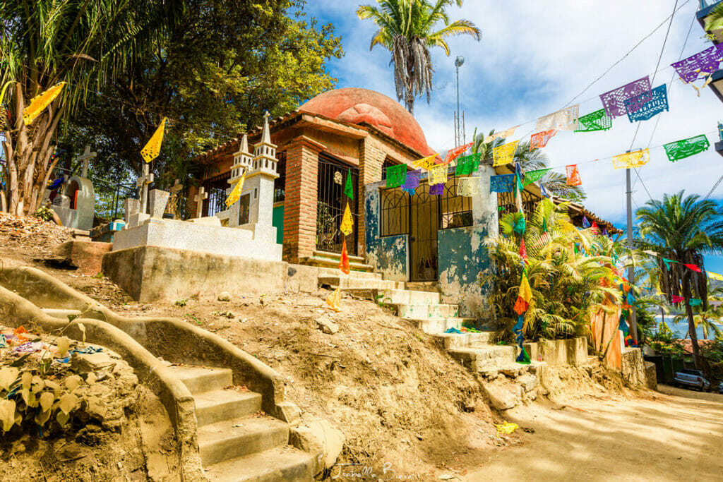 A small cemetery on a hillside with decorated graves and a brick building topped with a red dome. Colorful papel picado banners are strung across the scene, surrounded by lush greenery and palm trees.