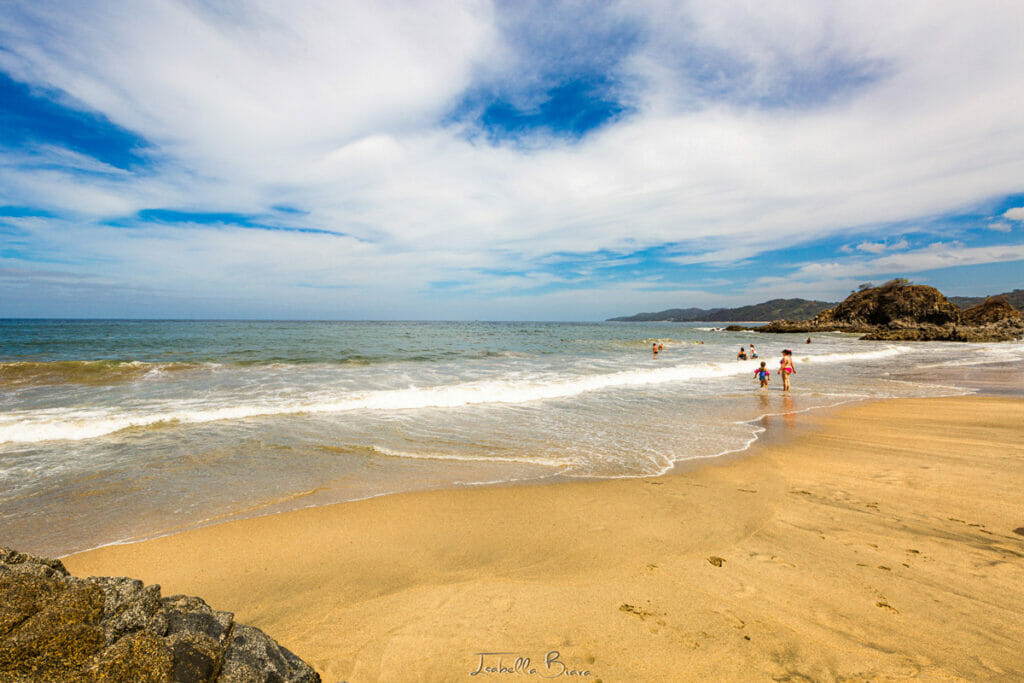 A golden sand beach with gentle waves rolling in under a partly cloudy blue sky. Several people are swimming or wading in the water near a rocky outcrop in the distance.