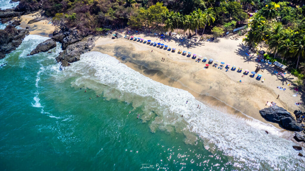 Aerial view of Playa de los Muertos in Sayulita, Mexico, showing turquoise waves gently crashing onto a sandy beach lined with colorful umbrellas and lounge chairs, surrounded by lush palm trees and rocky cliffs.