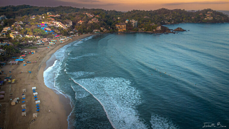 Aerial view of Sayulita's main beach with surfers in the waves, beach umbrellas lining the sand, and colorful hillside homes backing the bay.