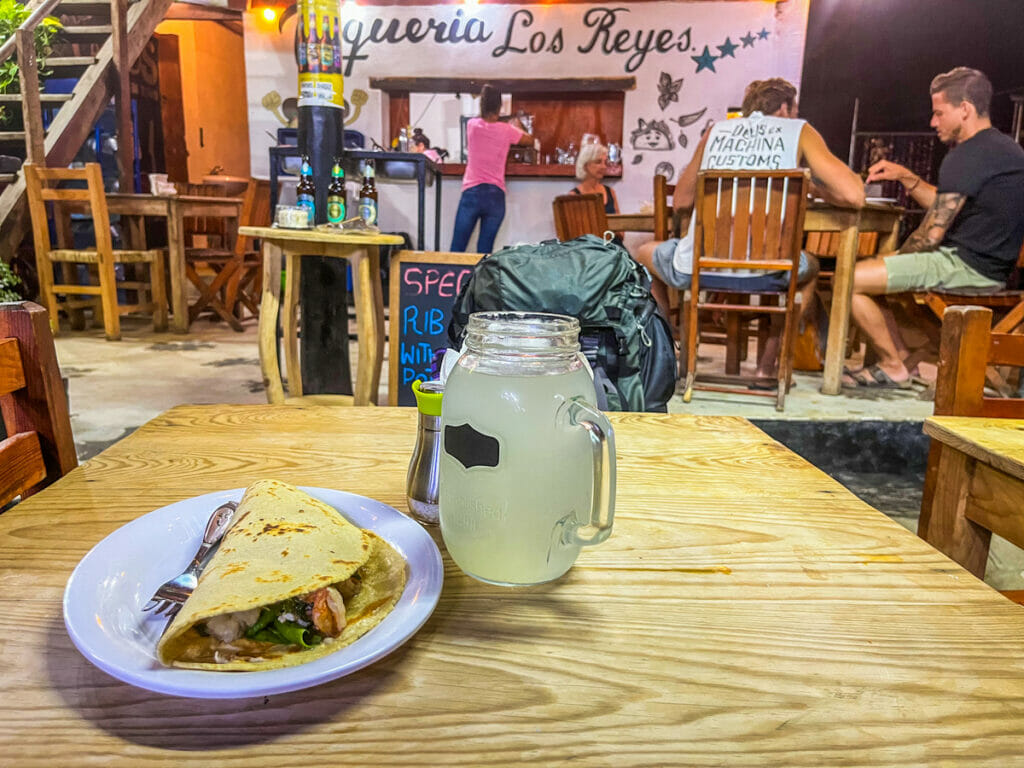 Close-up of a taco and a mason jar of lemonade on a wooden table, with the lively interior of Taquería Los Reyes in the background, including handwritten signs and customers dining.