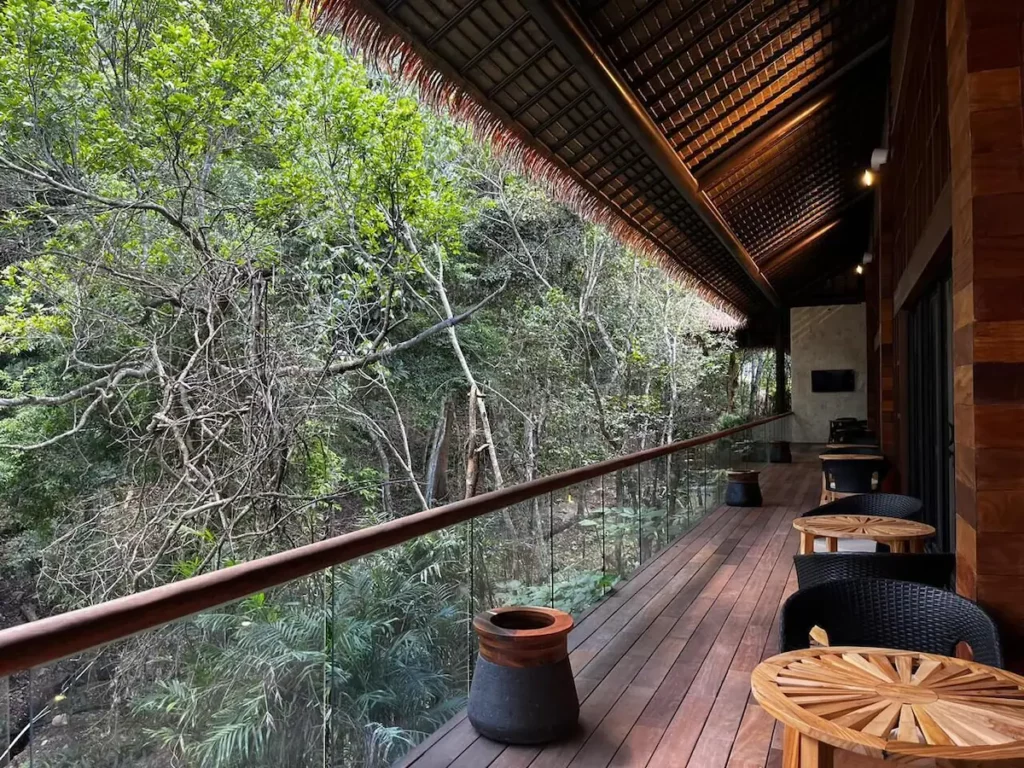 Wooden deck with woven chairs and tables, overlooking lush forest. Glass railing and thatched roof provide an open, inviting atmosphere in a tranquil setting.