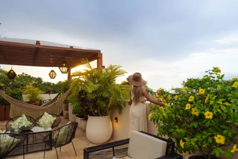 A person in a sun hat admires a rooftop garden with lush plants, hammock, and outdoor dining under a cloudy sky.