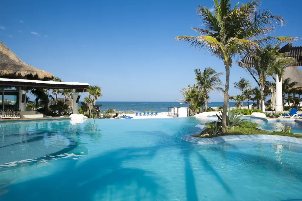 Tropical resort features pool, palm trees, and cabanas overlooking the ocean under clear skies, creating a serene vacation setting.
