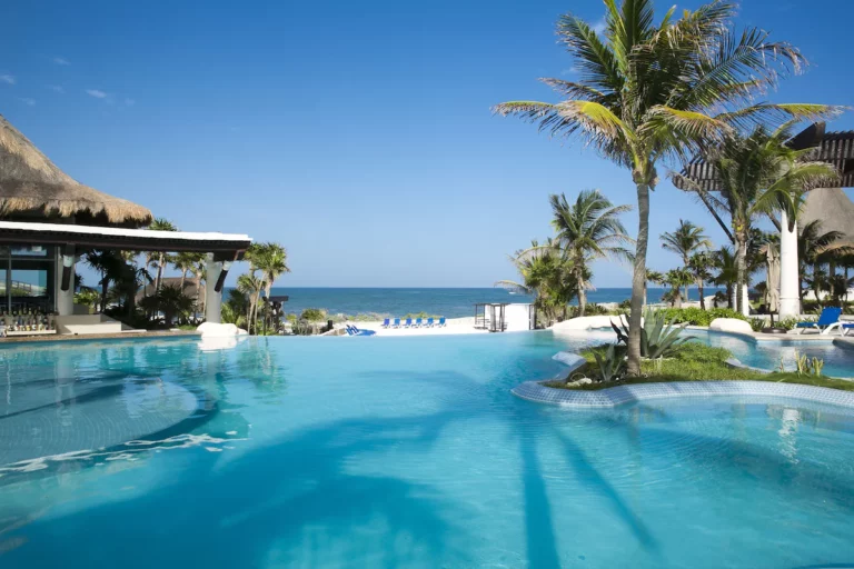 Tropical resort features pool, palm trees, and cabanas overlooking the ocean under clear skies, creating a serene vacation setting.