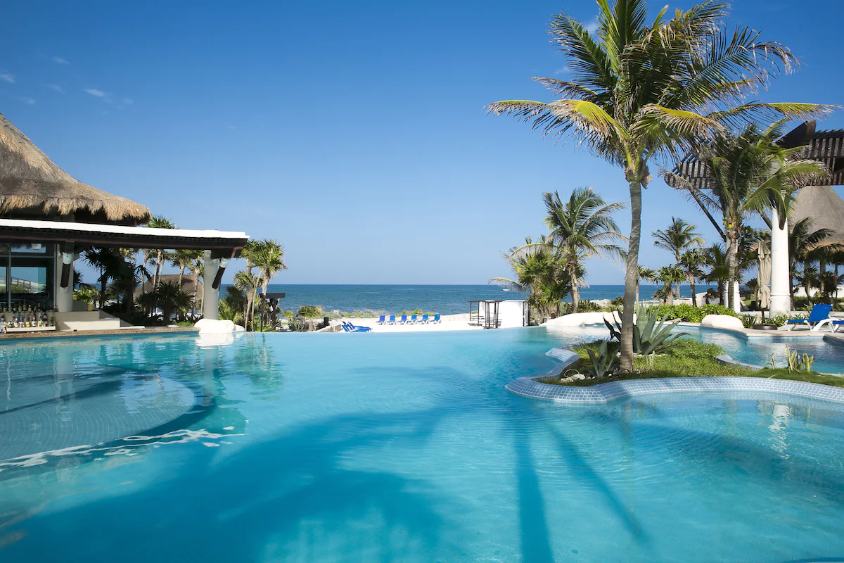 Tropical resort features pool, palm trees, and cabanas overlooking the ocean under clear skies, creating a serene vacation setting.