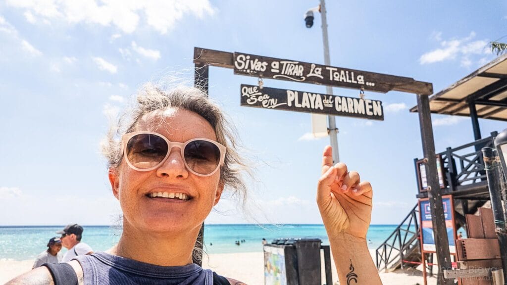 Smiling woman (me) wearing sunglasses pointing at a wooden sign that reads “Bienvenidos a Playa del Carmen” near a beach entrance, with the ocean and clear blue sky in the background.