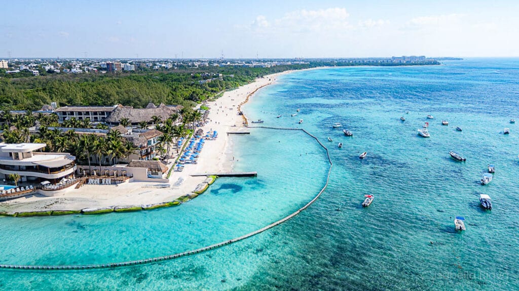 Aerial view of a vibrant beach with turquoise waters, boats, and shoreline buildings. Palm trees line sandy coast, with distant urban area visible.