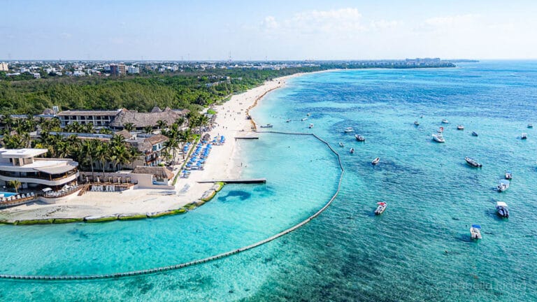 Aerial view of a vibrant beach with turquoise waters, boats, and shoreline buildings. Palm trees line sandy coast, with distant urban area visible.