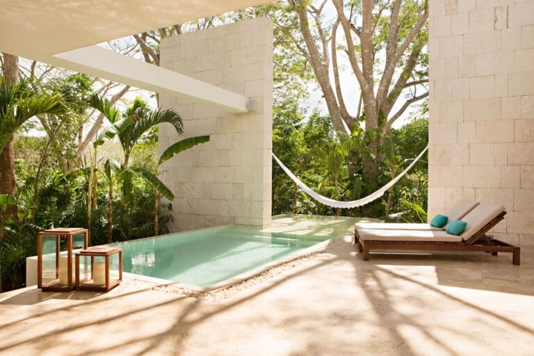 Outdoor pool area with a hammock, lounge chair, and nested tables, surrounded by lush tropical greenery and stone walls reminiscent of Yucatan haciendas.