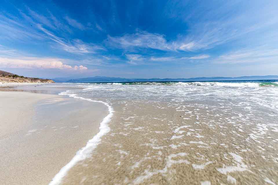 Shallow, crystal-clear waves gently curve onto the soft, pale sand of Playa Destiladeras, with the horizon stretching across a calm ocean under a streaked blue sky.
