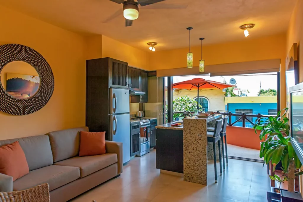 A compact and cheerful living space with a sofa, small kitchen, and breakfast bar, opening to a balcony with a bright orange umbrella and neighborhood views.