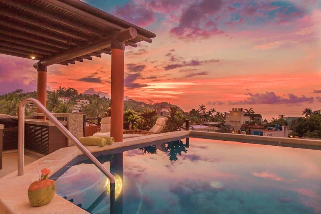 A rooftop infinity pool with a covered lounge area and deck chairs, set against a dreamy pink and purple sunset sky over a tropical coastal town.