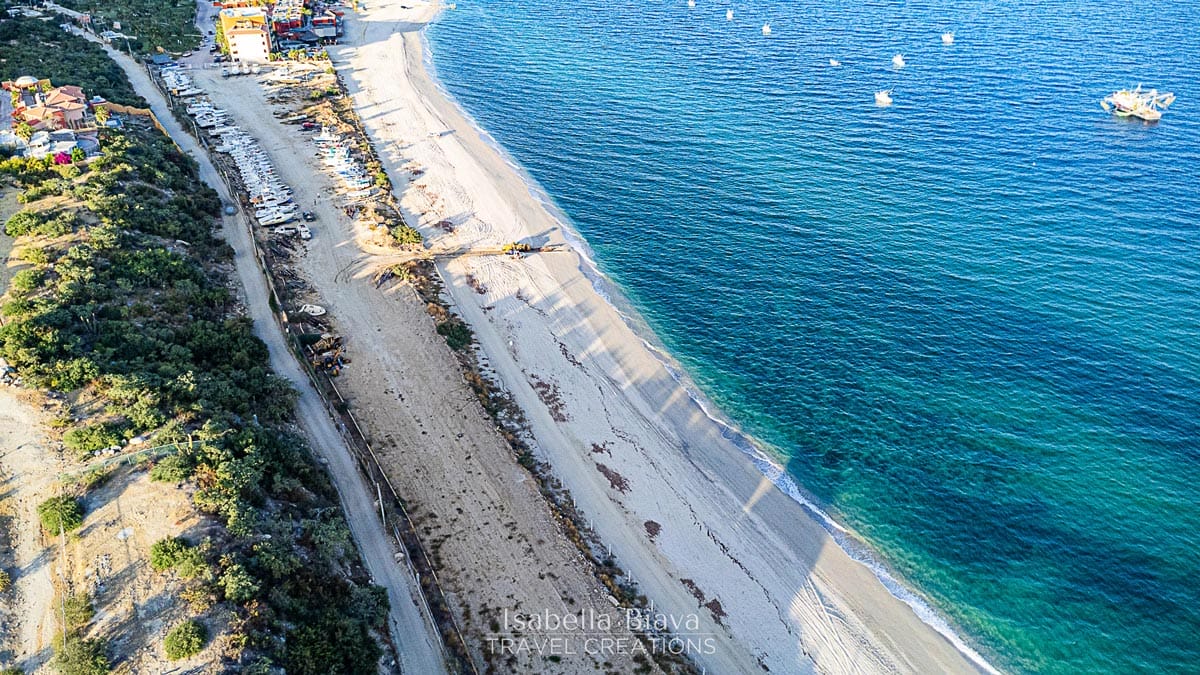 Aerial view of a sandy beach with clear blue waters, boats anchored offshore, and a small coastal town nearby, surrounded by greenery.