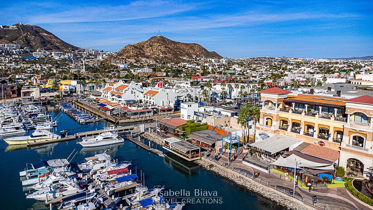 A vibrant marina with numerous boats docked alongside colorful buildings and shops, set against a backdrop of hilly terrain under a clear blue sky.