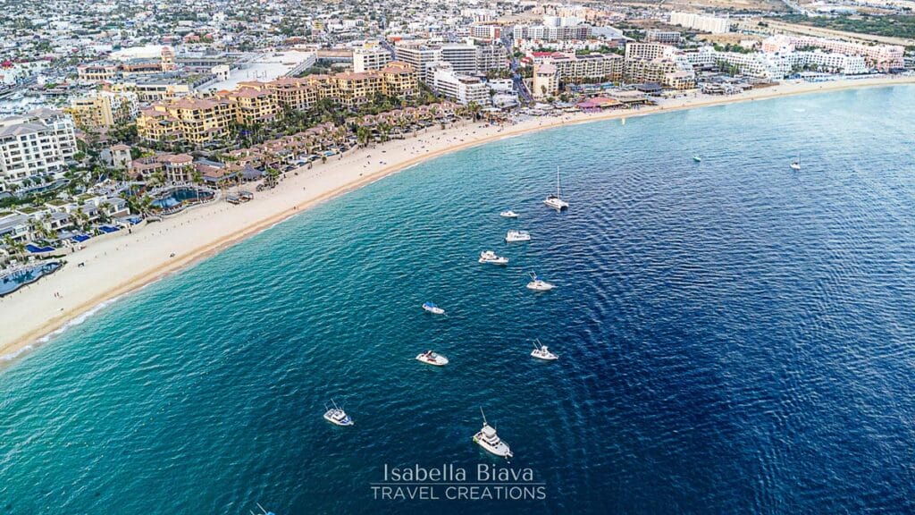 Aerial view of a vibrant beach with boats in the turquoise water, adjacent to a bustling coastal cityscape, under a sunny sky.