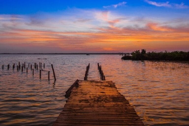 A wooden pier extends into a calm lake during a vibrant sunset, with scattered clouds painting the sky in hues of orange and blue.