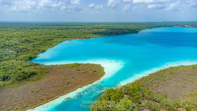 Aerial view of Bacalar Lagoon with its vibrant blue waters, surrounded by lush greenery and scattered red-roofed structures under a cloudy sky.