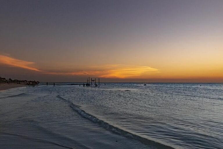 Sunset over a calm beach with gentle waves, silhouetted structures on the horizon, and a vast sky transitioning from orange to twilight.