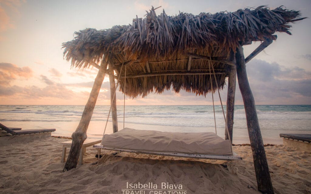 A rustic beach swing bed under a thatched roof sits on sand, overlooking a serene ocean at sunset, with soft clouds above.