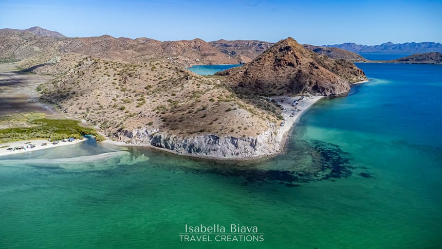 Aerial view of a scenic coastline with rocky hills, clear turquoise water, and a sandy beach, surrounded by lush vegetation under a bright blue sky.