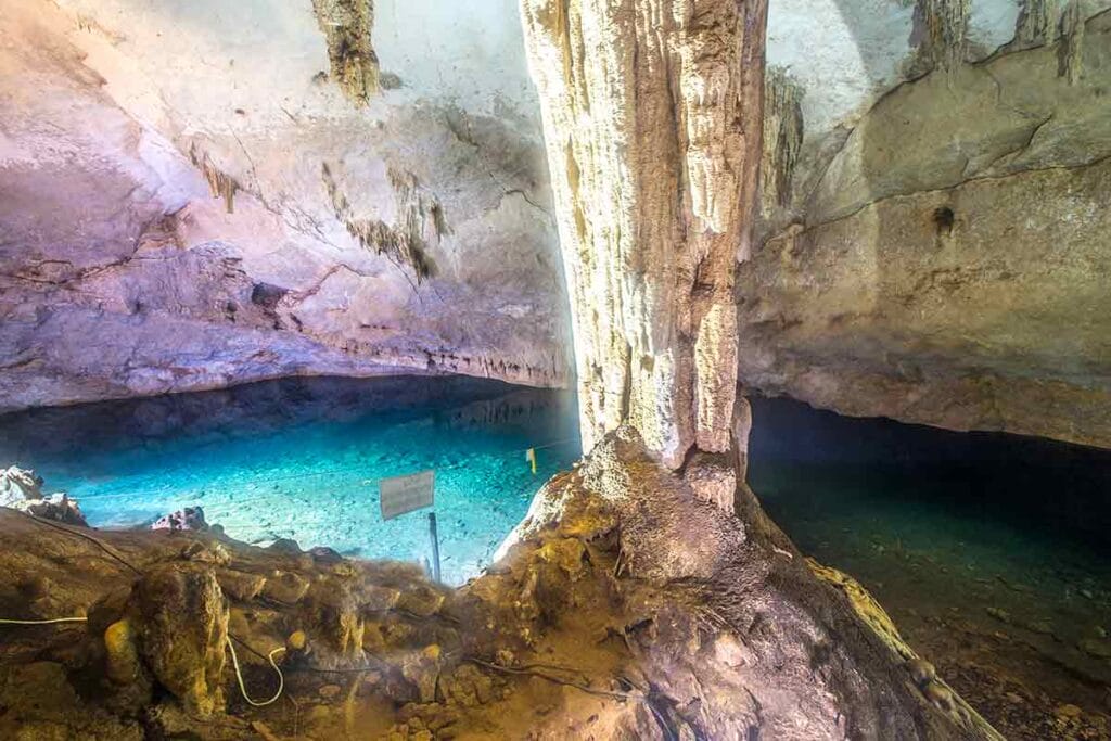 A stunning cave with a turquoise pool, stalactites, and stalagmites, illuminated by natural light filtering through the cave ceiling. No people present.