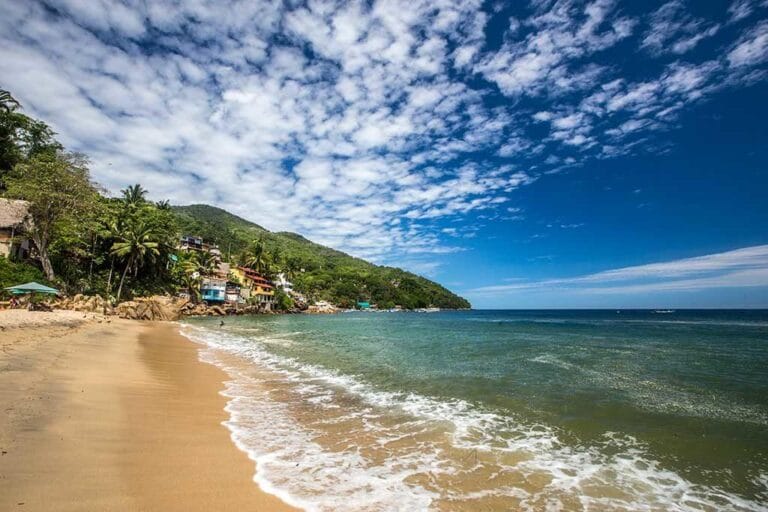 A secluded beach with lush greenery and colorful buildings along the shoreline, under a vast sky with scattered clouds.