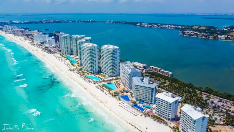 Aerial view of Cancún's beach lined with modern hotels, clear turquoise waters, and lush greenery. The cityscape stretches towards the distant horizon.
