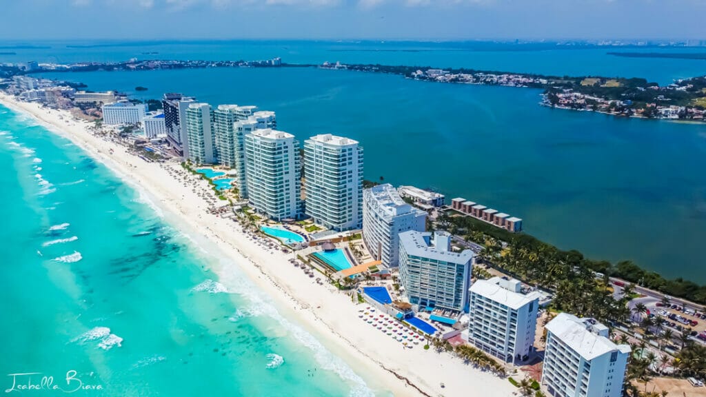 Aerial view of Cancún's beach lined with modern hotels, clear turquoise waters, and lush greenery. The cityscape stretches towards the distant horizon.