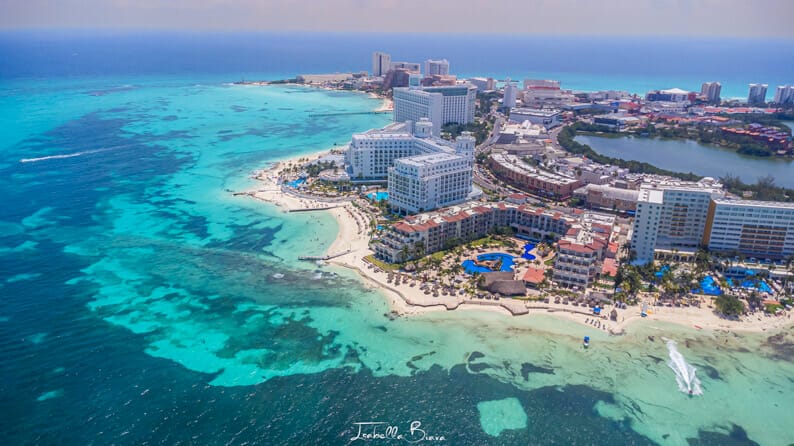 Aerial view of Cancun's coastline, featuring turquoise waters, sandy beaches, and numerous high-rise hotels against a vibrant blue sky.