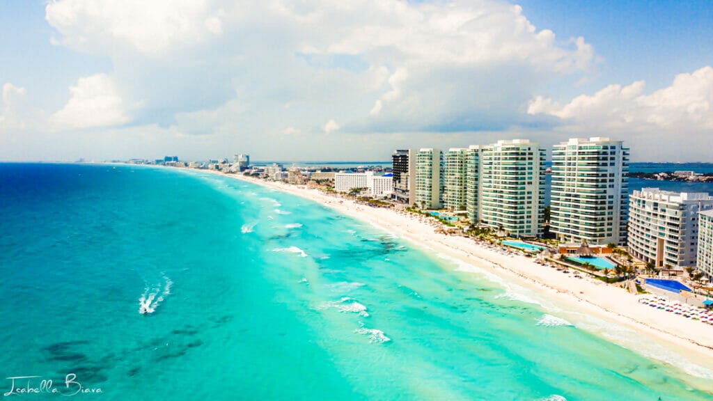 Aerial view of Cancun's coastline with turquoise waters, white sandy beach, and high-rise buildings. People enjoy activities in the vibrant beachfront area.