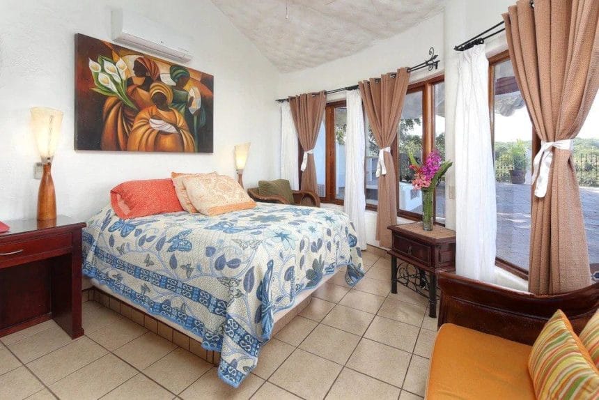 Bright bedroom with a queen-sized bed, colorful floral artwork, warm-toned bedding, and multiple windows with tan and white curtains letting in natural light.