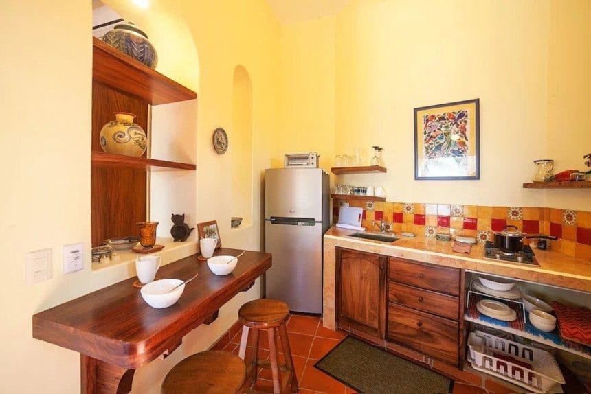 A compact kitchen with warm yellow walls, colorful tile backsplash, wooden cabinetry, and a small breakfast bar set for three.