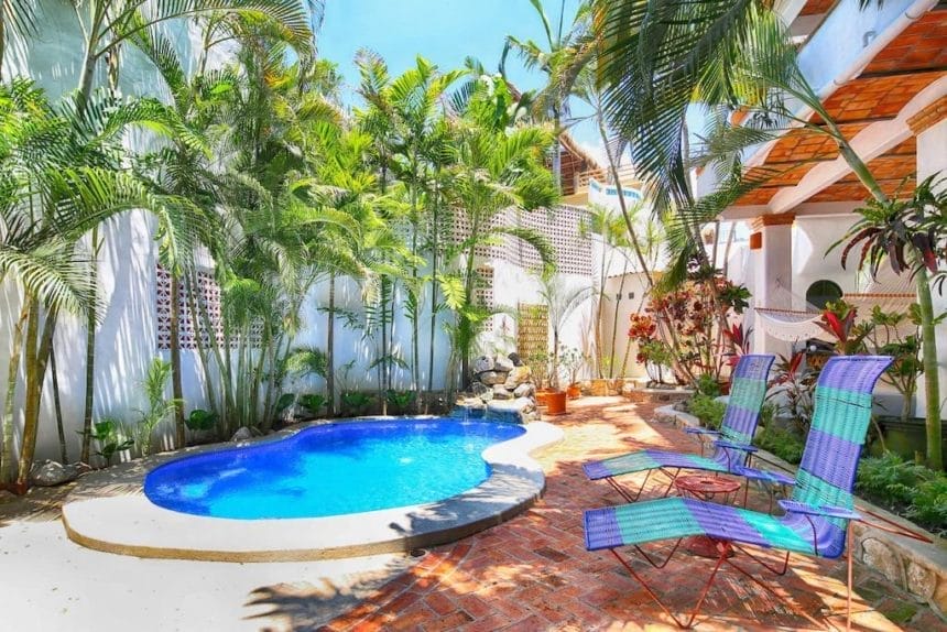 Small private pool nestled in a walled courtyard, surrounded by tropical plants, red brick flooring, and colorful poolside lounge chairs.