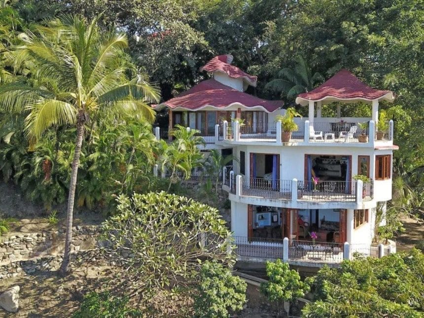 Multi-level white villa with red wave-like roofing, balconies on each floor, surrounded by dense tropical greenery and palm trees.