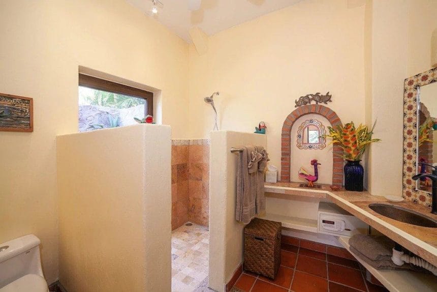 A cozy bathroom with cream walls, a walk-in shower lined with terracotta tiles, and vibrant Mexican-style decor including painted figurines and a floral vase.