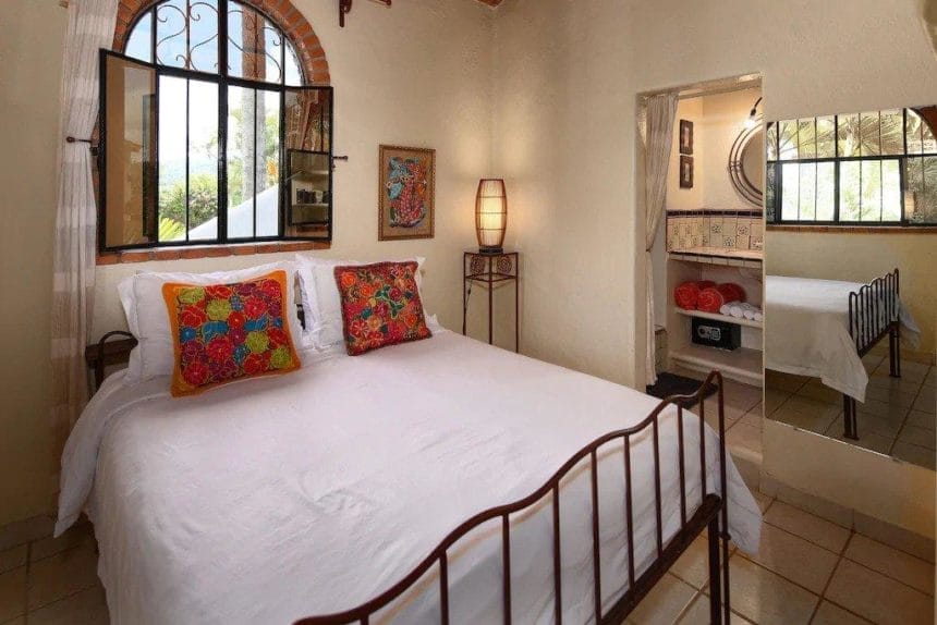 Cozy bedroom with a wrought iron bed, bright white linens, colorful embroidered pillows, and a small ensuite bathroom with vibrant tiles.